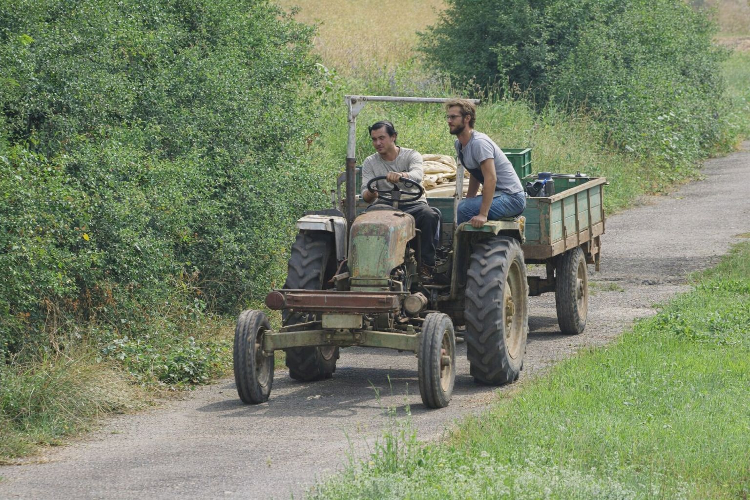 Två män på en äldre grön traktor med släp på en grusväg vid en familjegård.